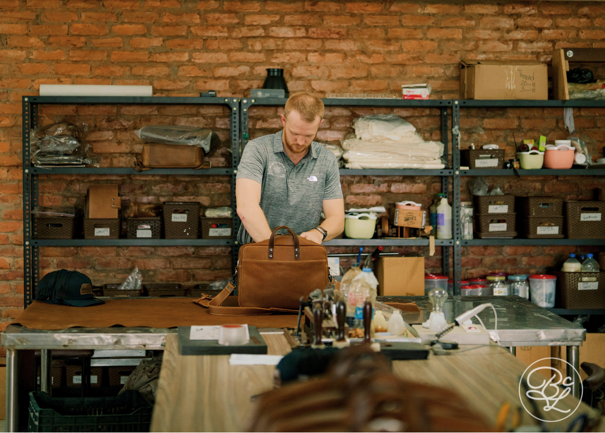 A man working at a leather crafting table inside a workshop with a brick wall background, handling a brown leather bag. Shelves with tools and materials are visible behind him.